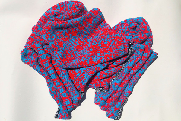An image of a machine knit artwork by Kate Geck called Knitting with GANs. Generative Adversairial Networks were trained on vintage knitting machine patterns to create new designs that were knit into a textile on a domestic knitting machine. It is a vibrant red and blue optic pattern.